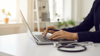 A close-up side-angle photograph of a healthcare professional in smart black business attire typing on a laptop and holding a pen. A stethoscope rests on the white desk beside the laptop. The background shows a bright, airy office space with shelving, plants, and natural light from large windows, conveying a professional private clinic governance setting.