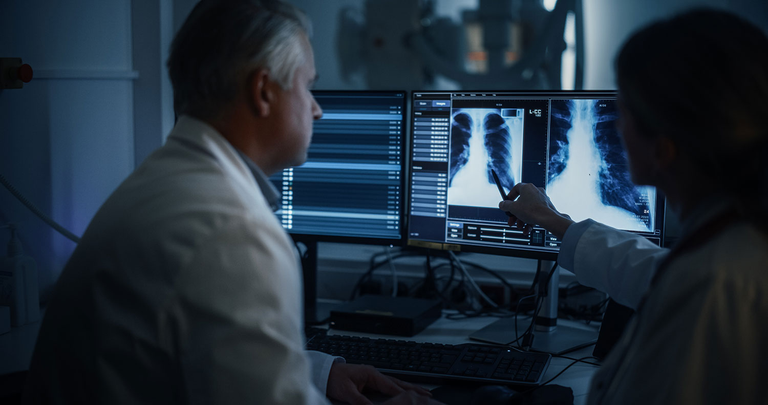 Clinical photo showing two healthcare professionals in a dimly lit room reviewing chest X-rays displayed on computer screens, with one person pointing at the images.