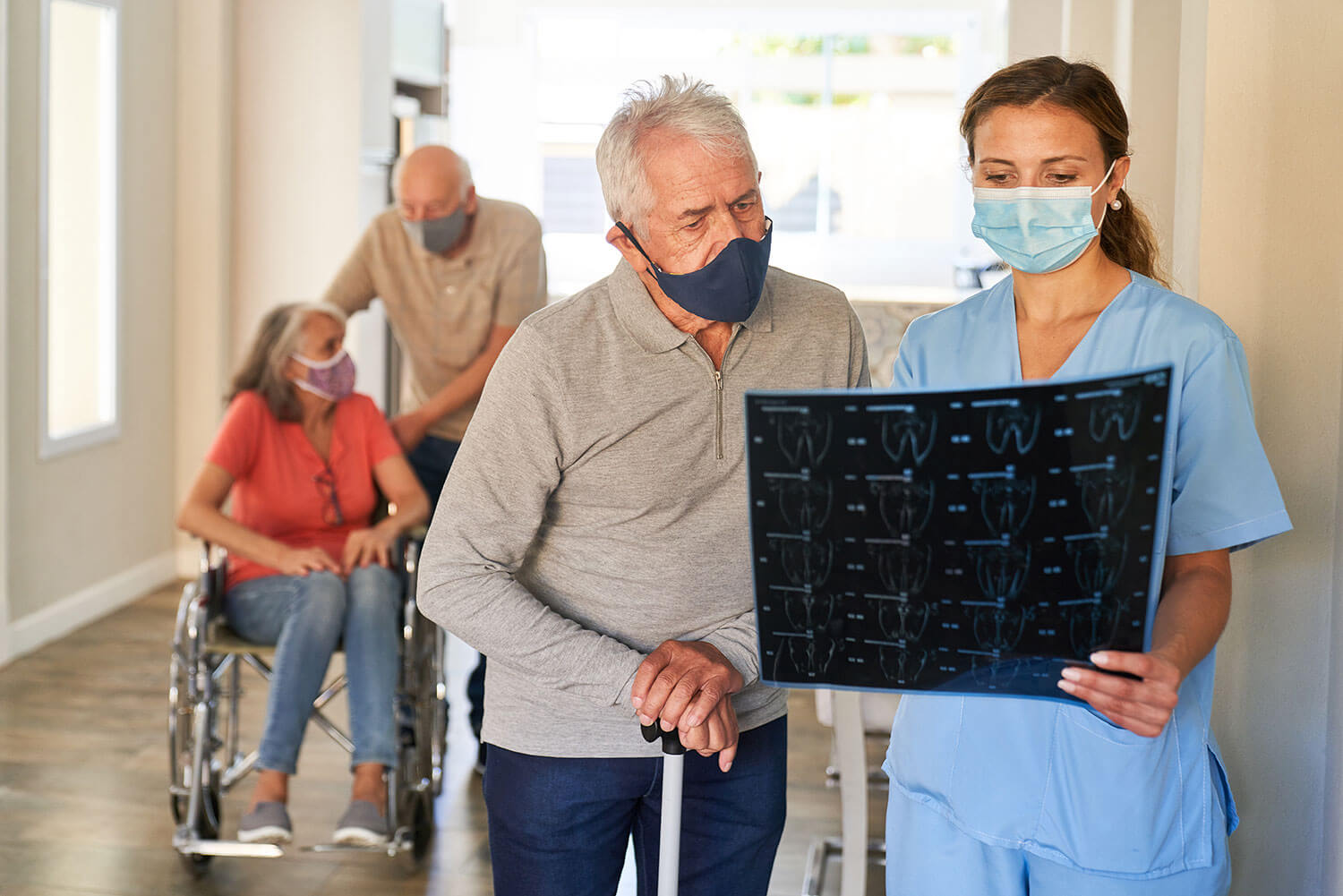 Photo of a masked nurse holding a medical scan and explaining it to an older patient using a walking stick, with other residents and carers in the background.