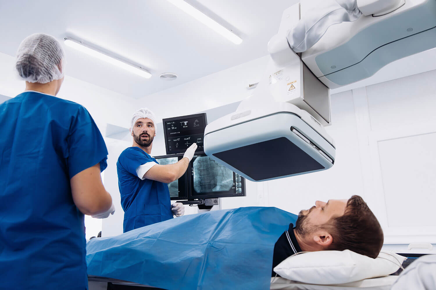 Clinical photo showing a patient lying on a bed while two healthcare workers in scrubs review images on monitors and operate overhead medical imaging equipment.